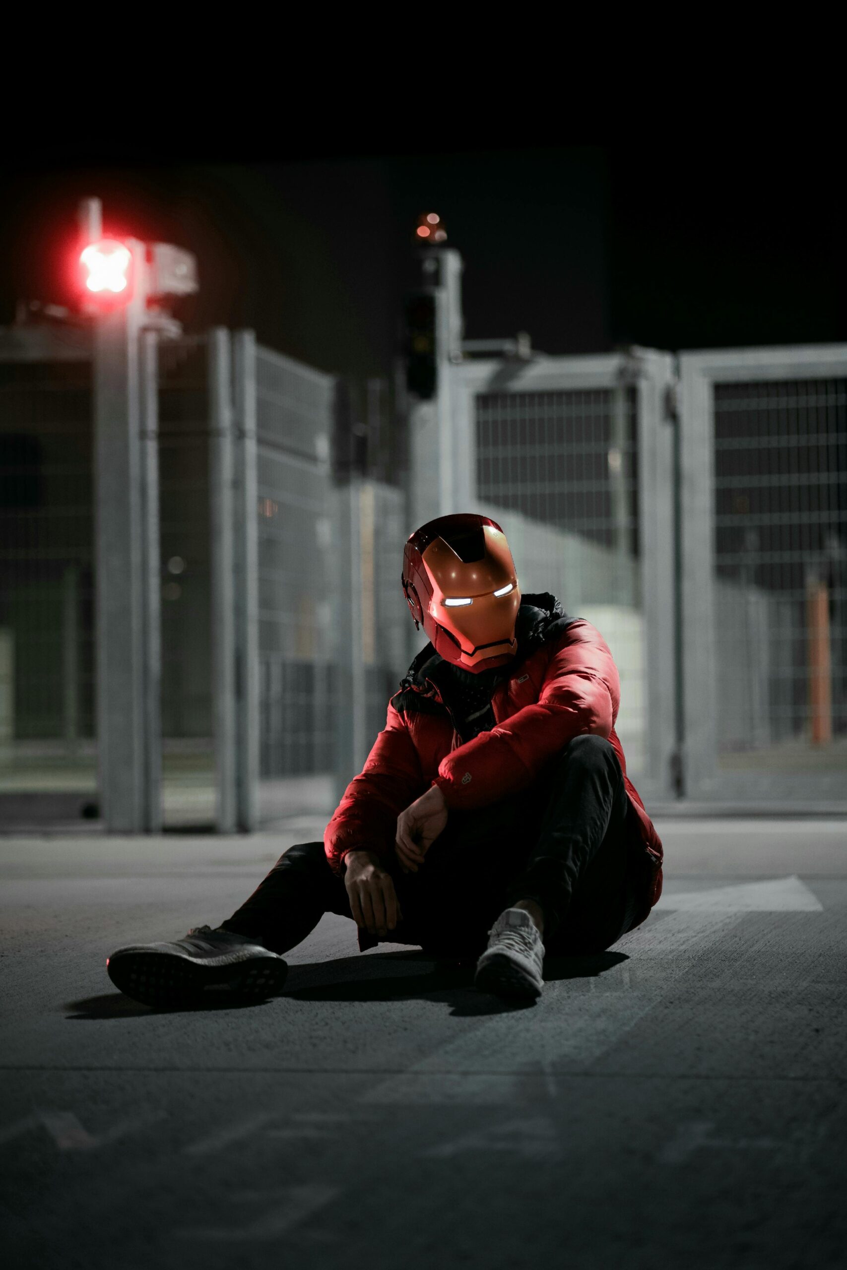 A person wearing an Iron Man helmet sits on the ground at night in an urban setting.